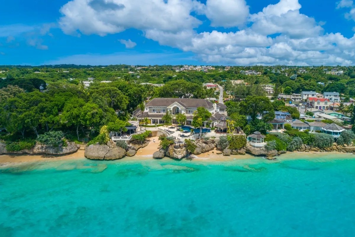 Aerial view of a large waterfront mansion with pool above a turquoise reef shoreline