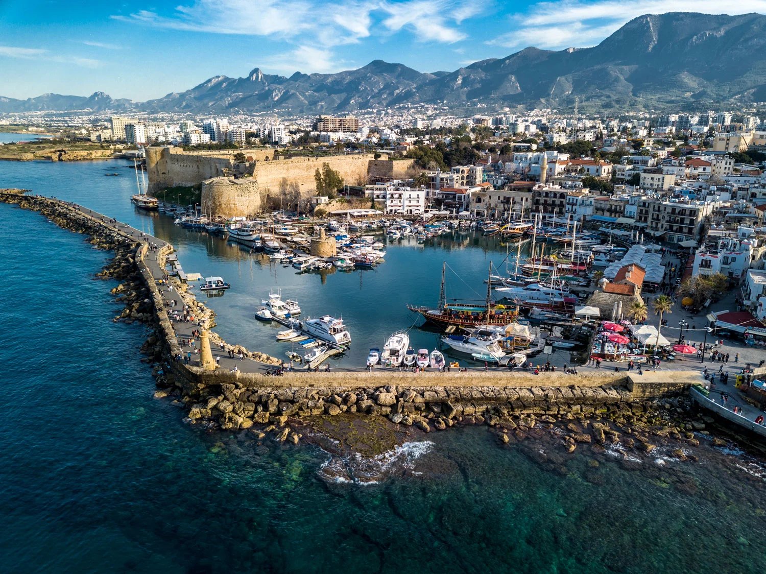 Aerial view of a stone castle and curved breakwater sheltering a boat-filled harbour