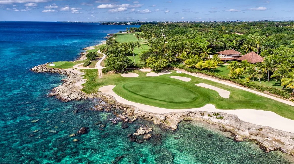 Aerial view of a coastal golf green and bunkers beside clear shallow water.