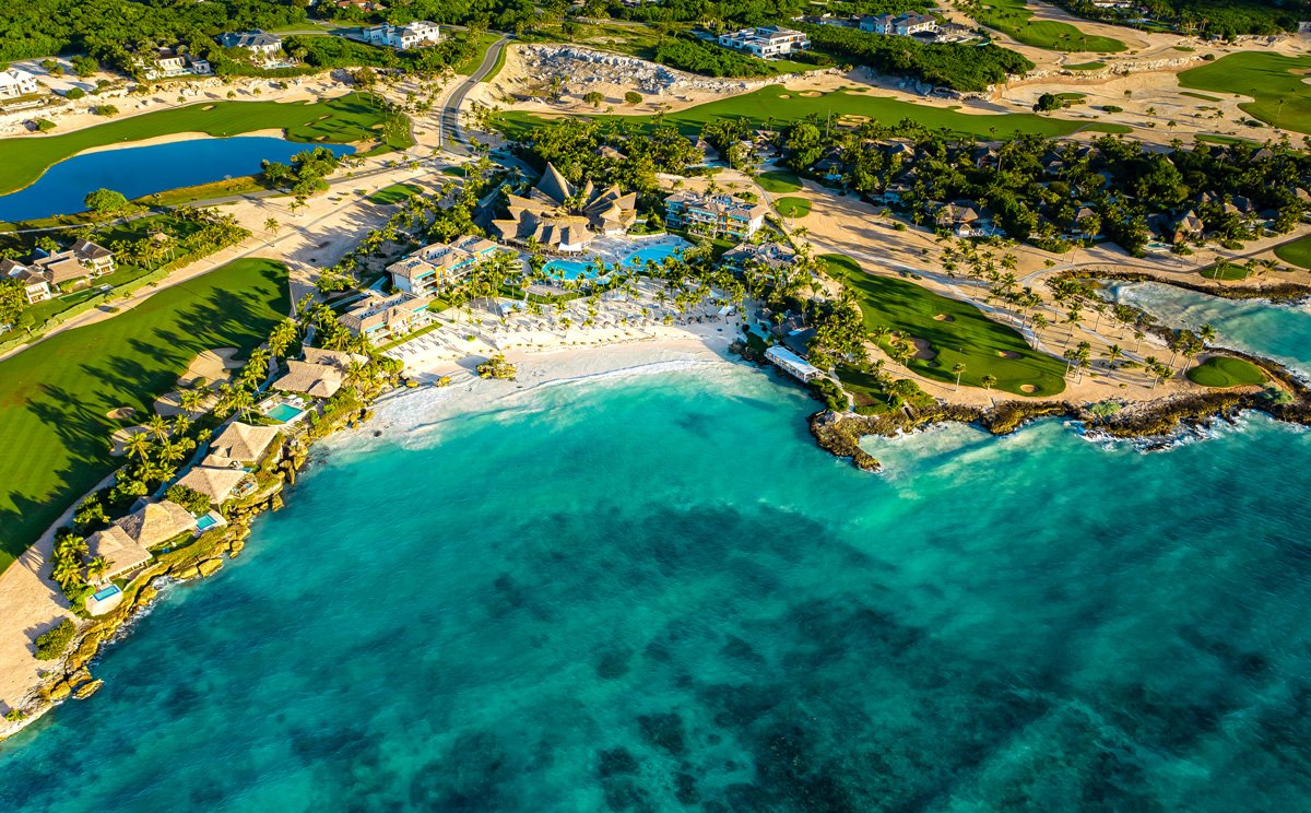 Aerial view of a beachfront resort and golf greens beside turquoise shallows.