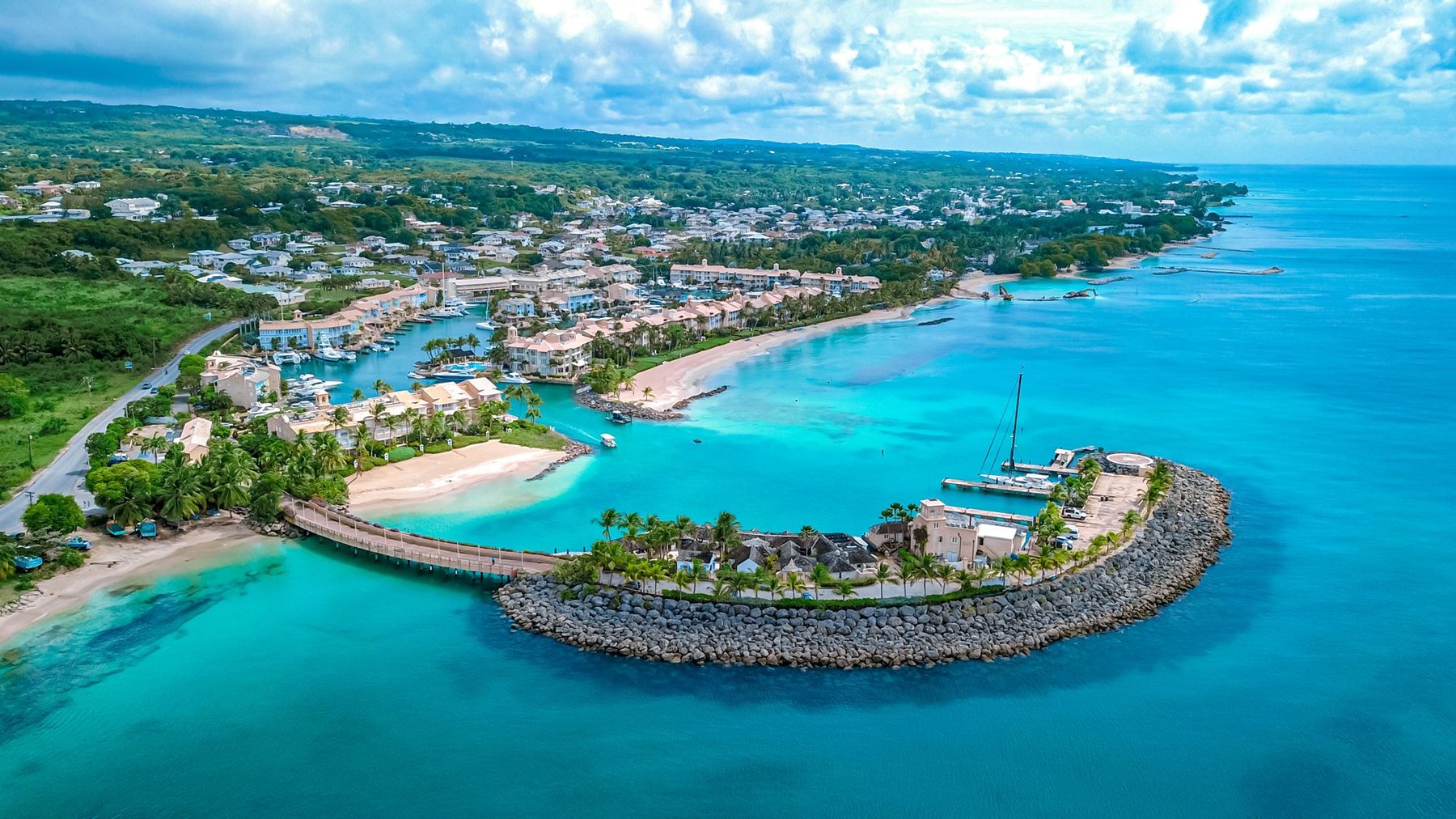 Aerial view of a curved rock breakwater, marina and low-rise resort town by turquoise water