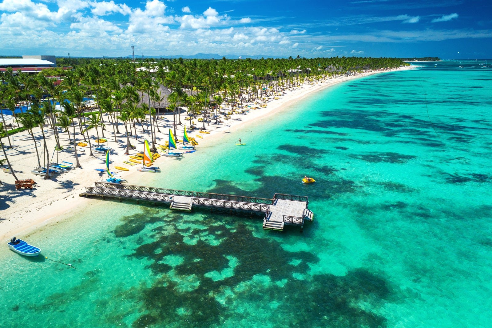 Aerial view of a wooden pier reaching over turquoise water from a palm beach.