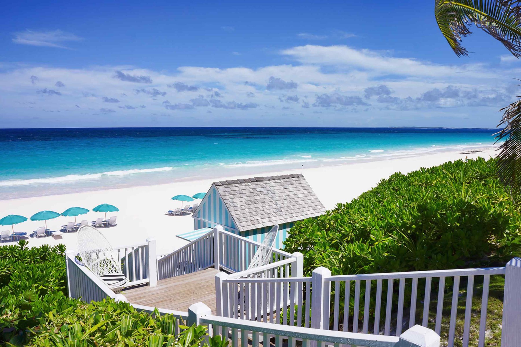 A white timber deck with hanging chairs above pale sand, a striped cabana, turquoise umbrellas and loungers, open ocean beyond