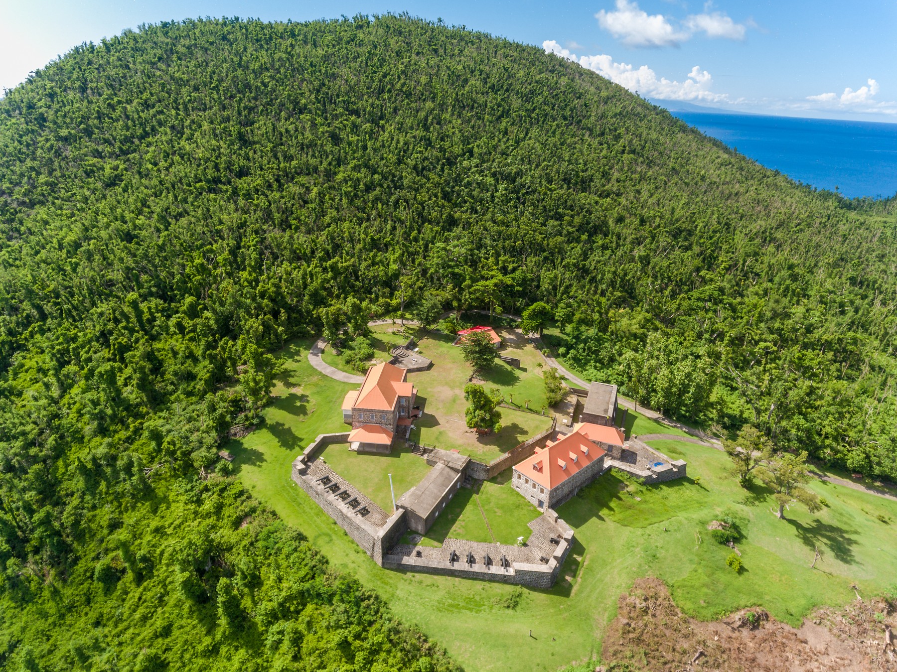 An aerial view of Fort Shirley at the Cabrits, its stone barracks and orange roofs set in cleared ground surrounded by dense rainforest above the sea
