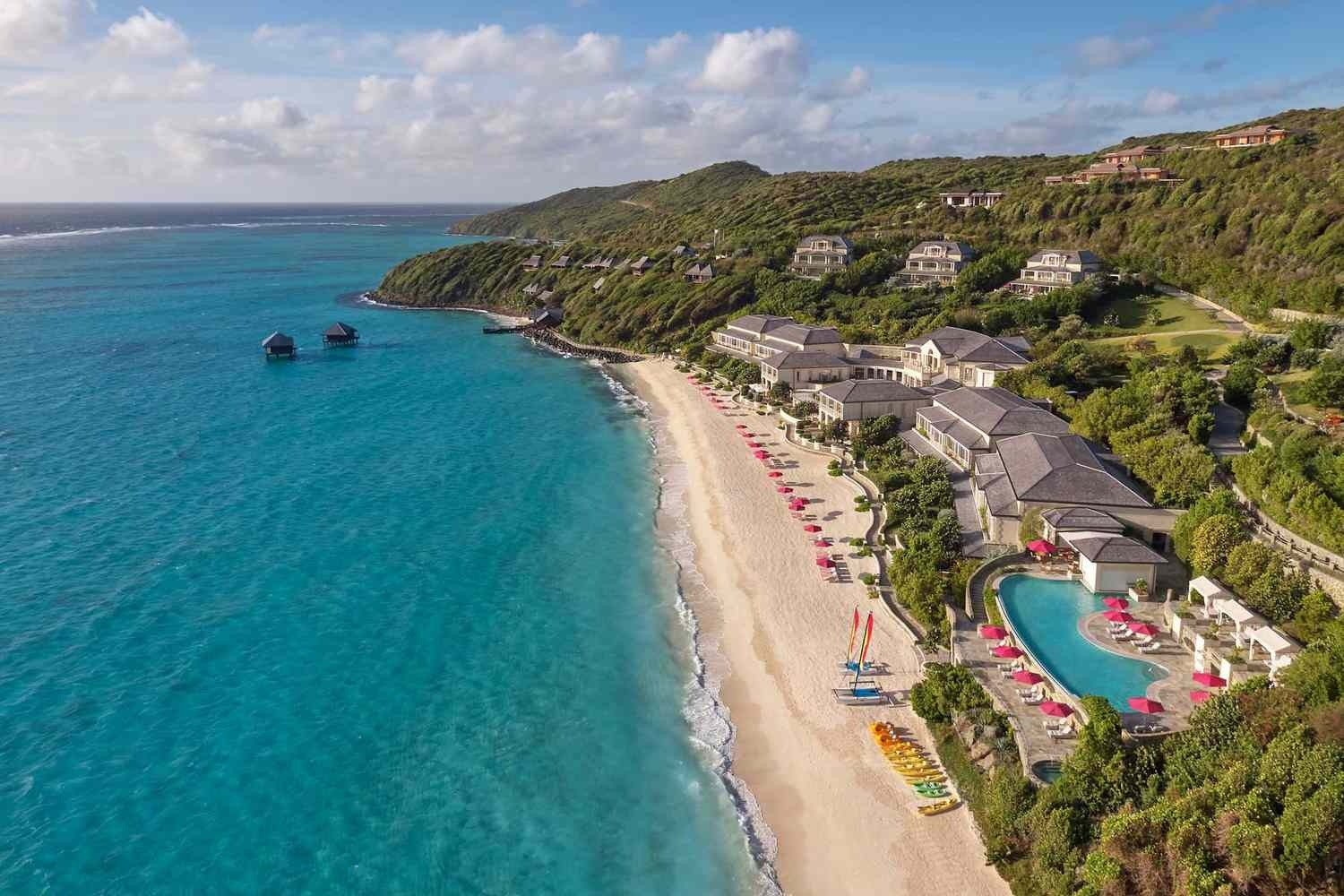 Resort buildings and villas stepping up a green hillside above a curving pale beach, with a pool terrace and umbrellas at one end