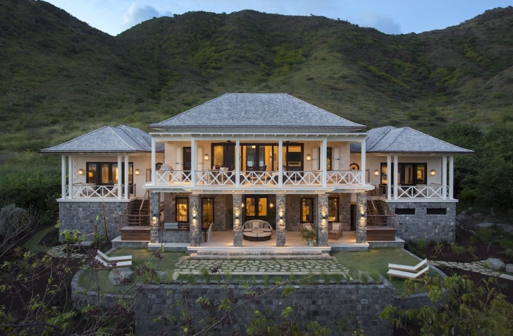 Stone-and-shingle villa lit at dusk against green hillside at Christophe Harbour, St Kitts