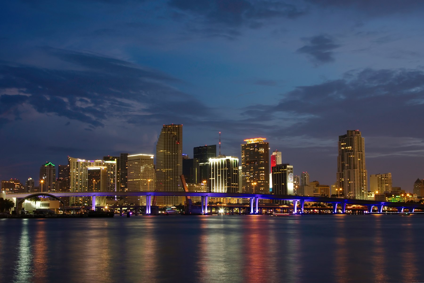 Downtown skyline and a violet-lit causeway bridge reflected in calm water at dusk