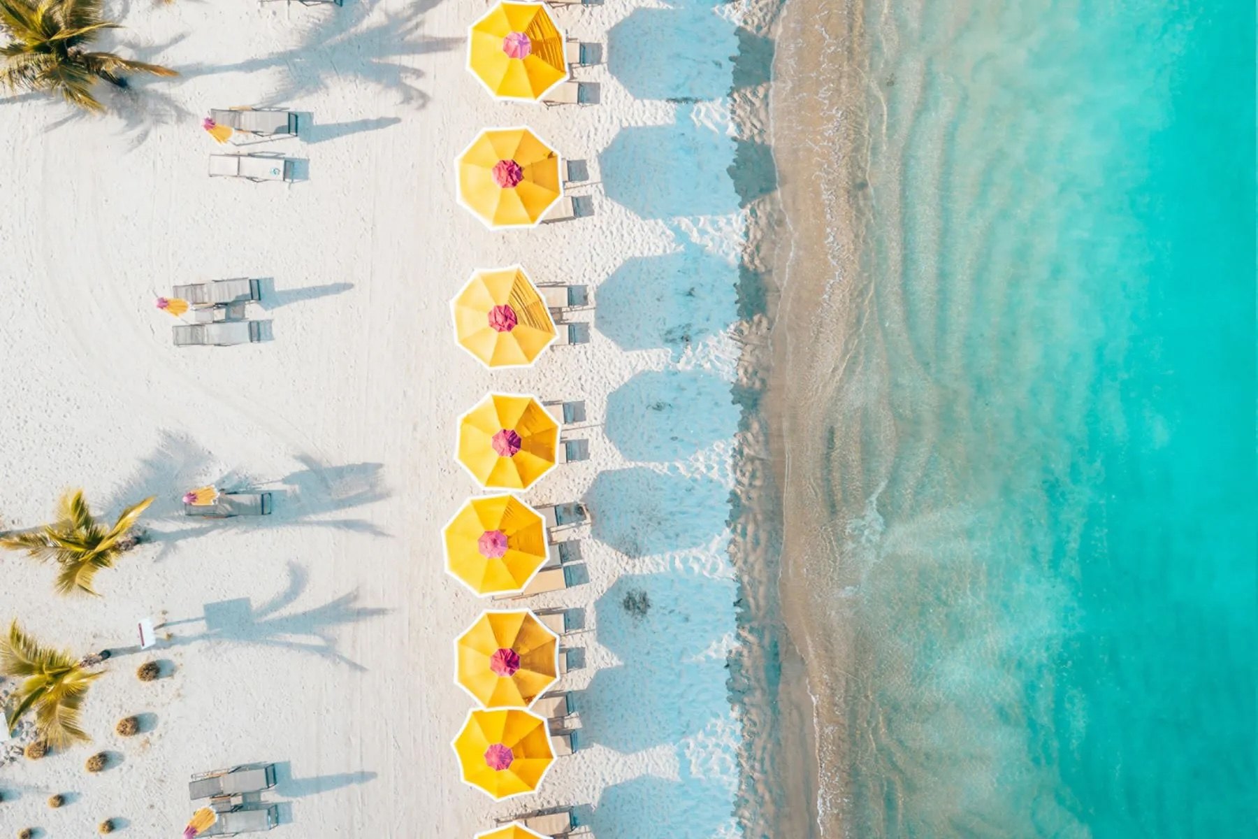 Overhead view of yellow beach umbrellas and loungers set in a line along white sand beside clear turquoise shallows