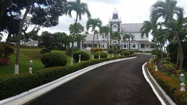 Curved driveway lined with clipped hedges leading to a white colonial government house