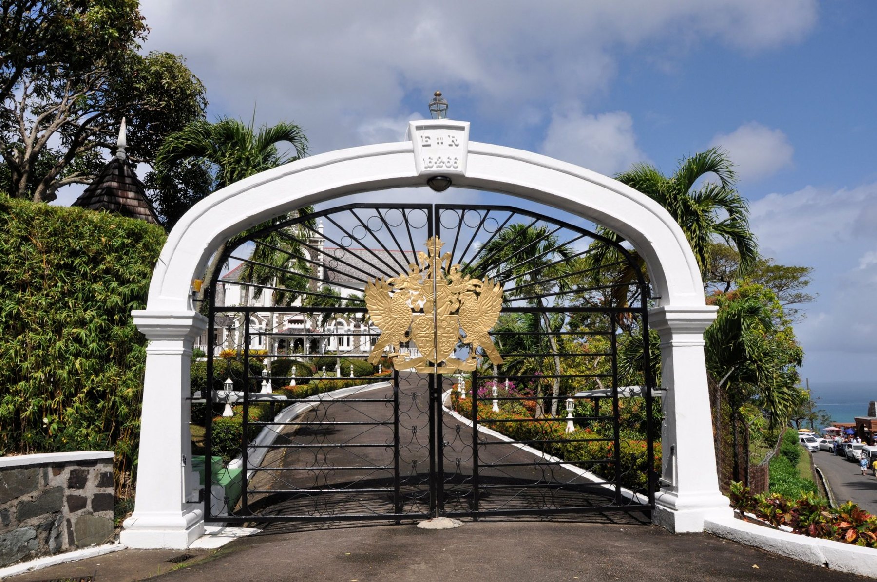 Whitewashed arch and black wrought-iron gates with a gilded crest, drive curving to a white house