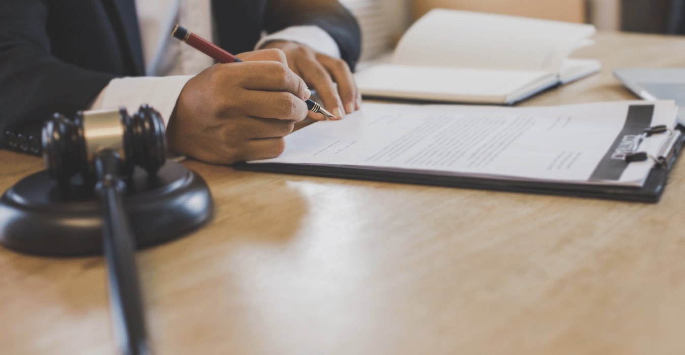 A hand signs a printed document on a desk beside a black wooden gavel and notebook
