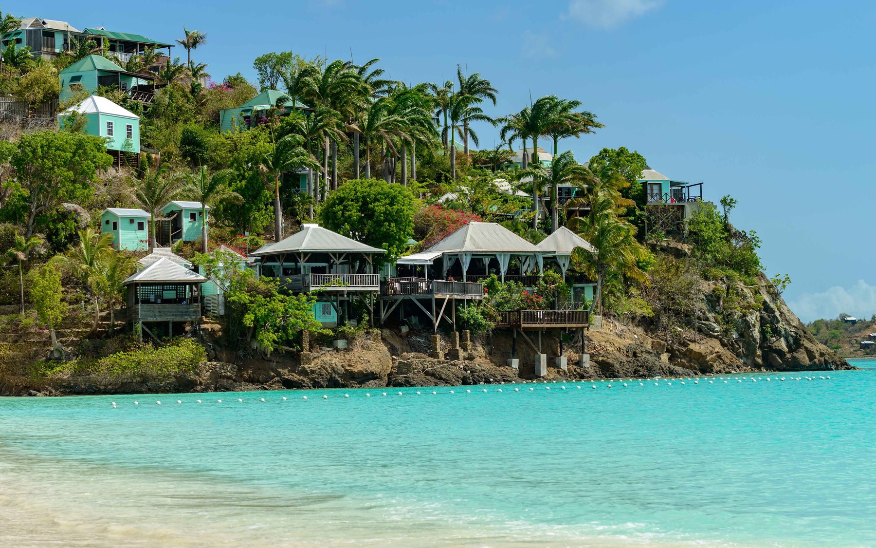 Pastel timber cottages raised on stilts above a clear turquoise Caribbean bay, framed by coconut palms