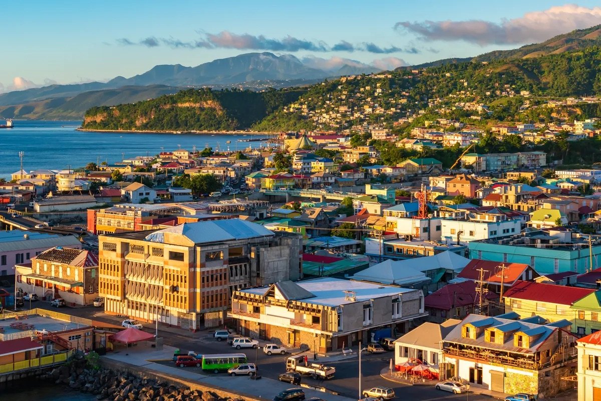 Roseau, Dominica at golden hour — dense, brightly roofed town buildings packed along the seafront beneath steep rainforest mountains