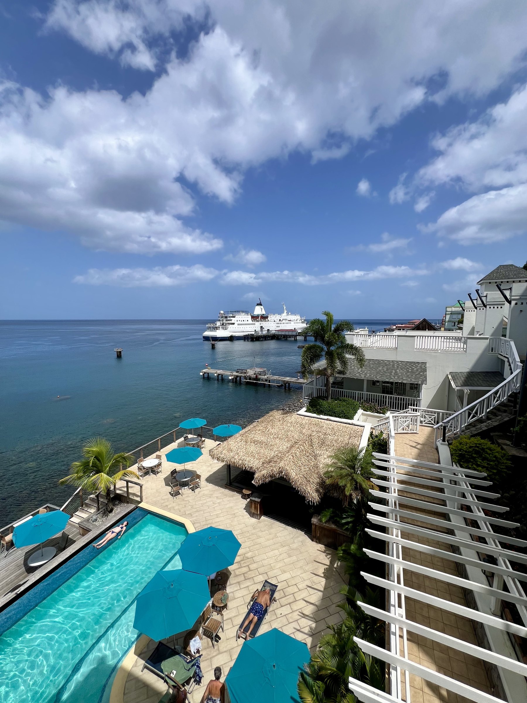 Hotel pool with blue parasols above a pier where a ship is docked.