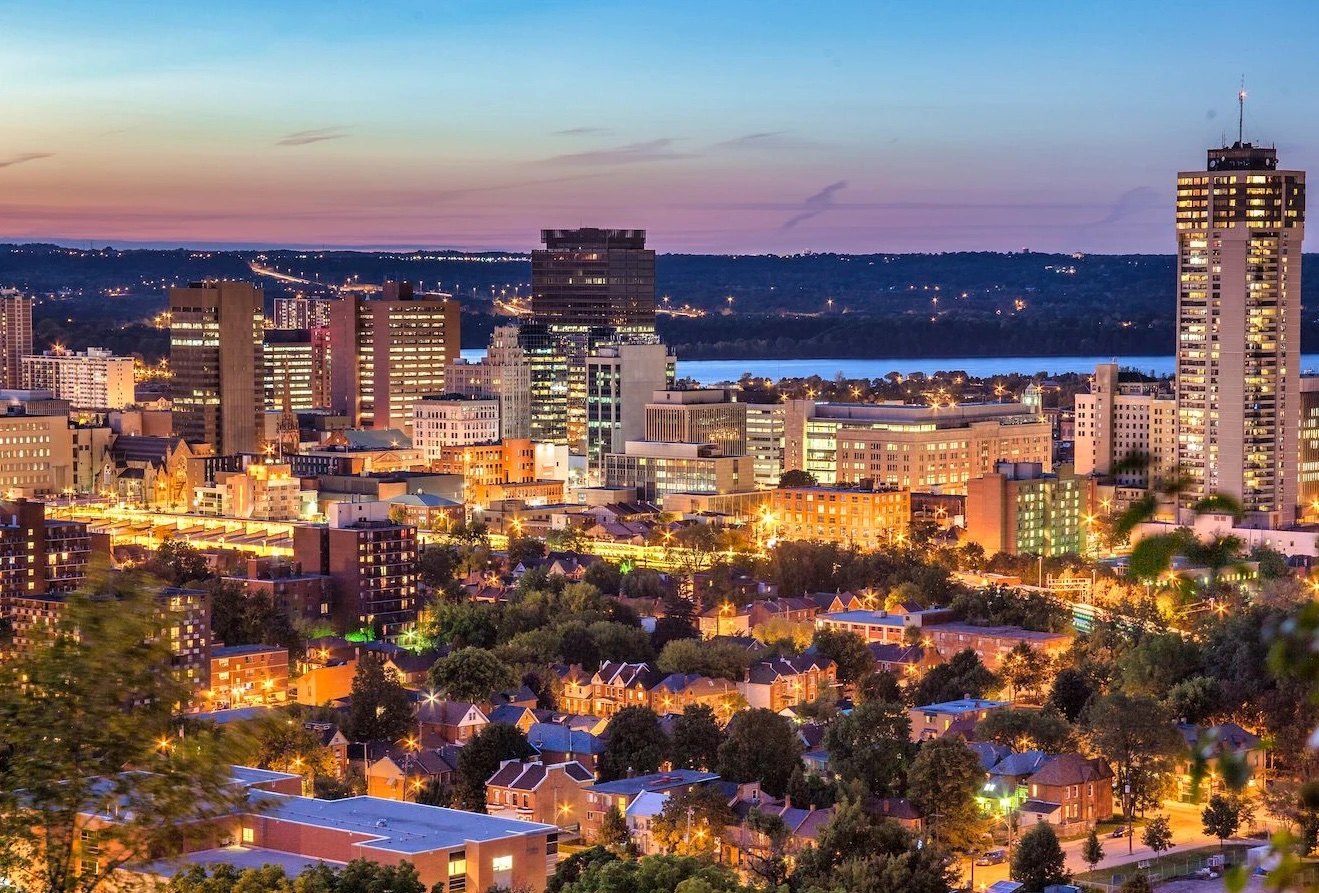 Downtown towers and tree-lined residential streets glowing at dusk above a distant bay
