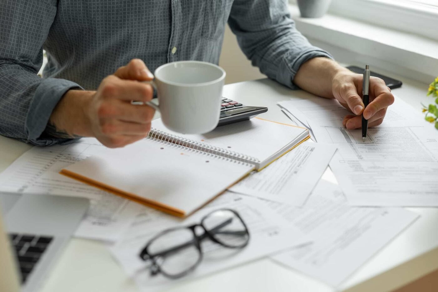 A man at a desk working through tax forms and paperwork, with a calculator, notebook and coffee cup