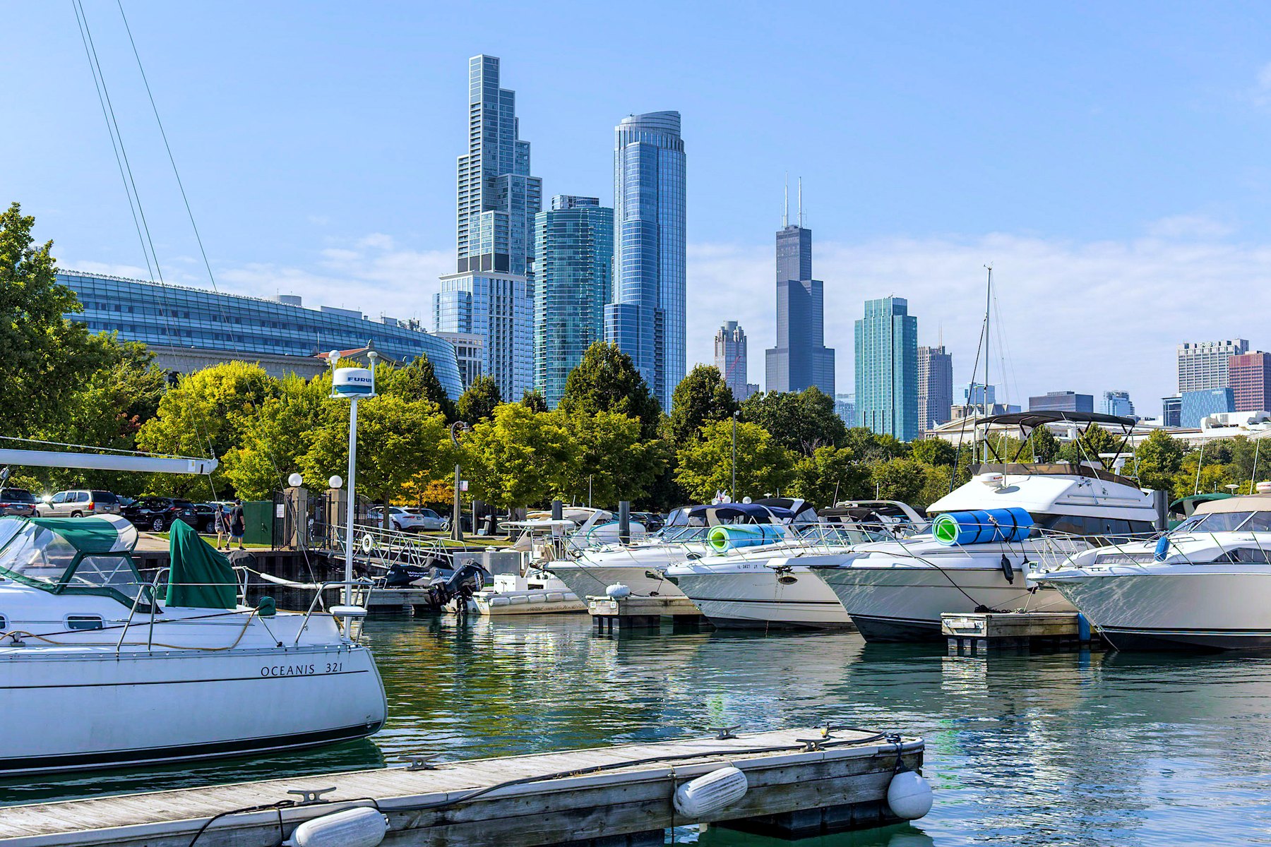 White motor yachts and a sailboat moored at a city marina below a downtown skyline