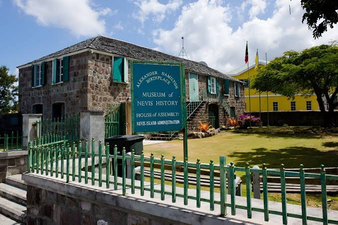 Stone building with green shutters signed House of Assembly, Nevis Island Administration, behind green railings