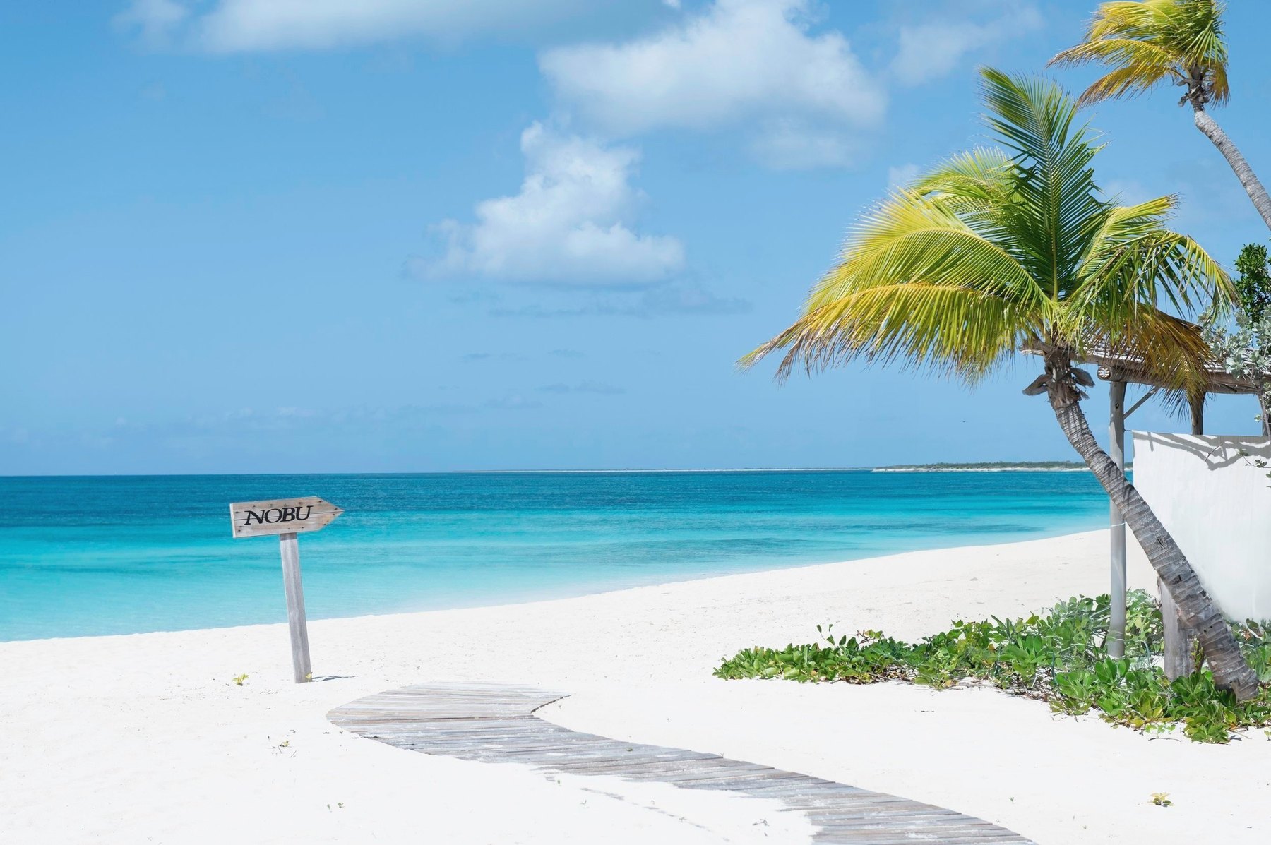 Weathered Nobu signpost beside a boardwalk on Barbuda's white sand and turquoise shallows