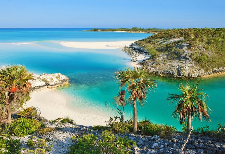 Palms on a rocky bluff above a turquoise cove, with a white sandbar curving out towards a low green cay