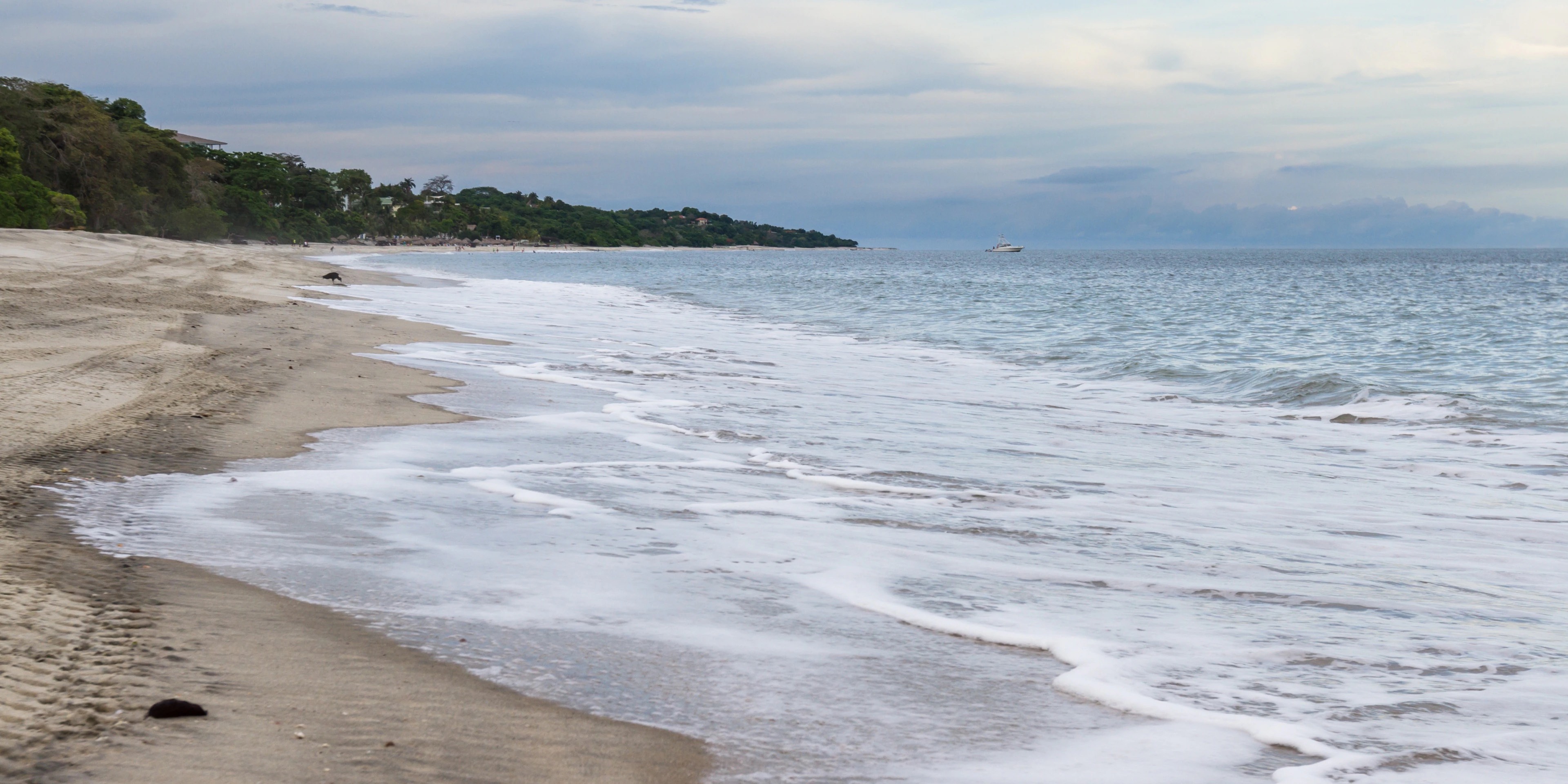 Wide grey-sand beach at dusk on Panama's Pacific coast, with surf running ashore, a forested headland of low-rise buildings beyond, and a small boat anchored offshore