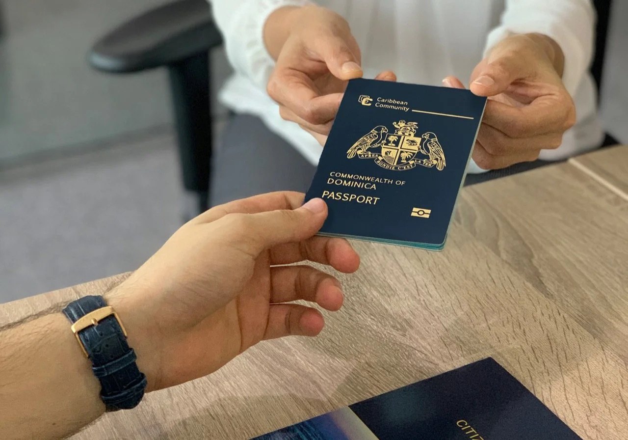 One hand passes a navy passport to another hand across a wooden desk