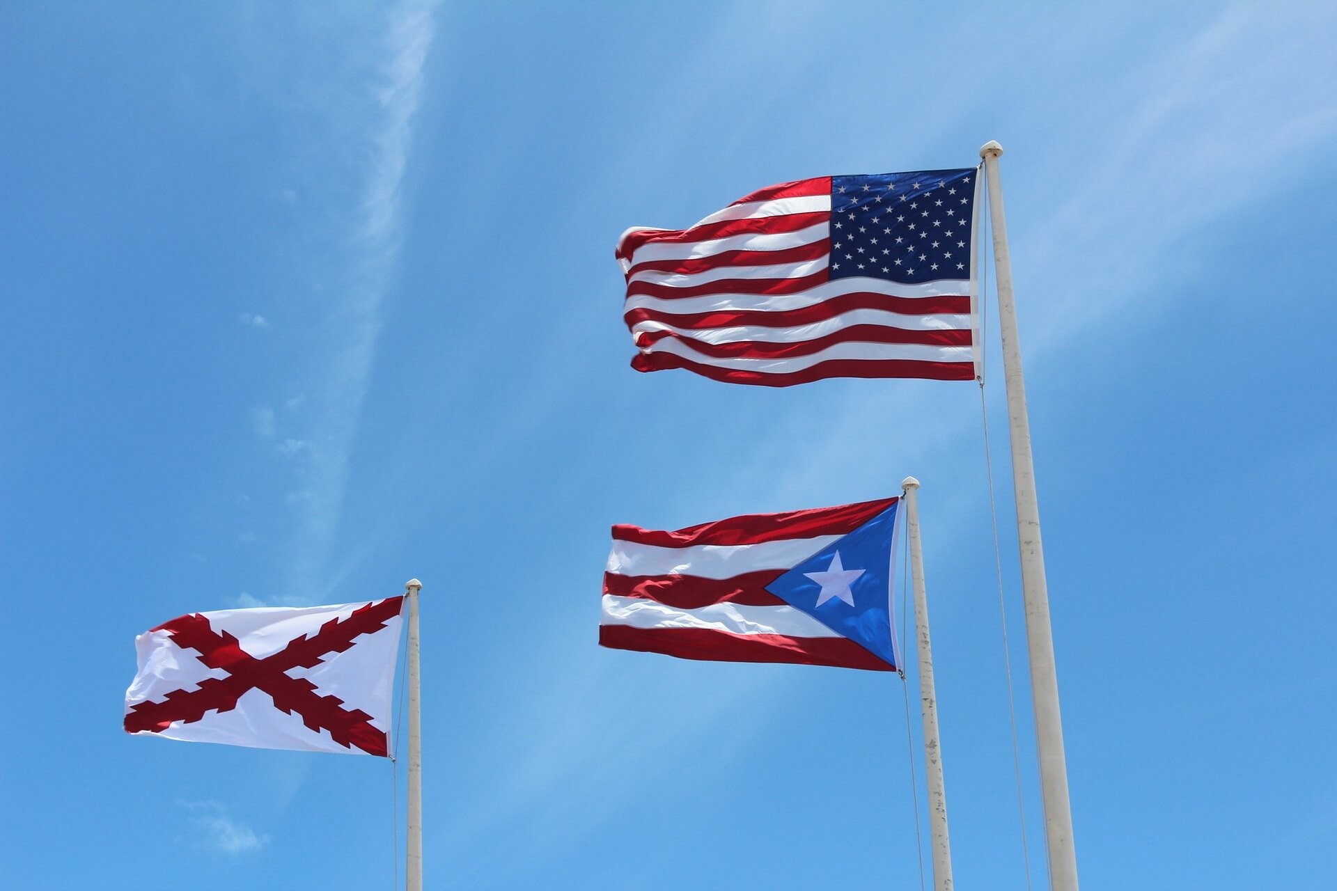 The flag of the United States, the flag of Puerto Rico and the Spanish Cross of Burgundy flying from three white poles against a clear blue sky