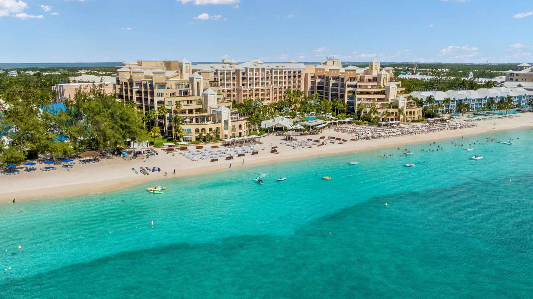 The Ritz-Carlton, Grand Cayman seen from the water — a sand-coloured resort behind the white sand and turquoise shallows of Seven Mile Beach