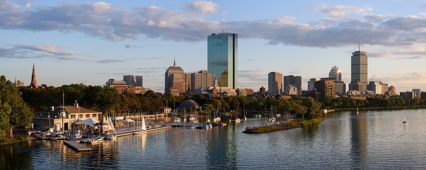 Sailboats moored beside a riverside boathouse with a downtown skyline in golden light