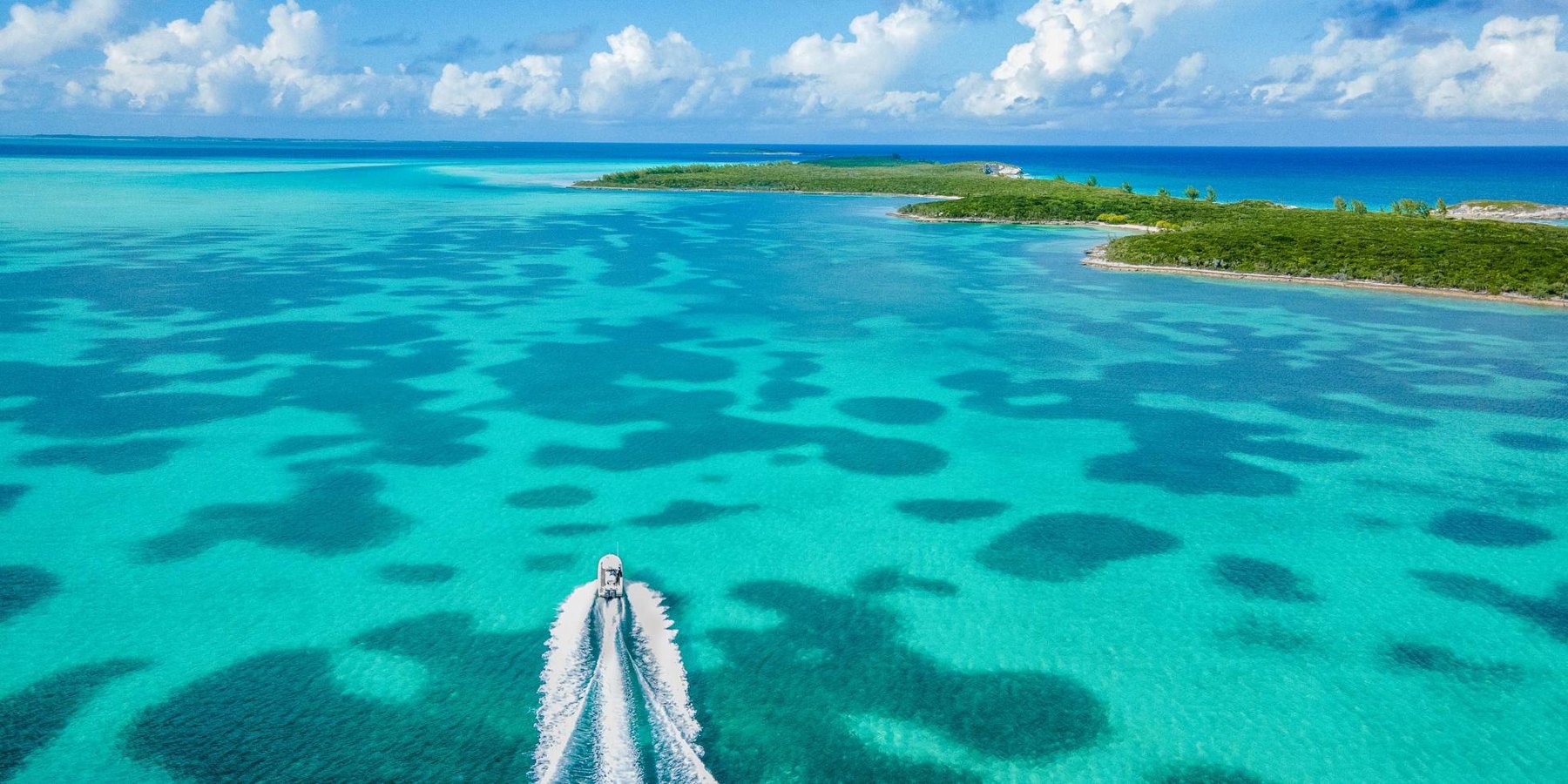 Small motor yacht speeding across clear turquoise shallows toward a low green island