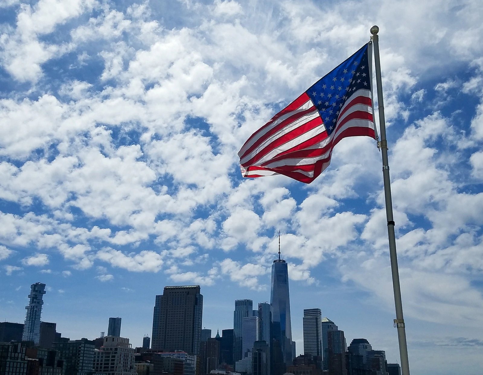 Stars and stripes flag flying above a downtown skyline under scattered white clouds