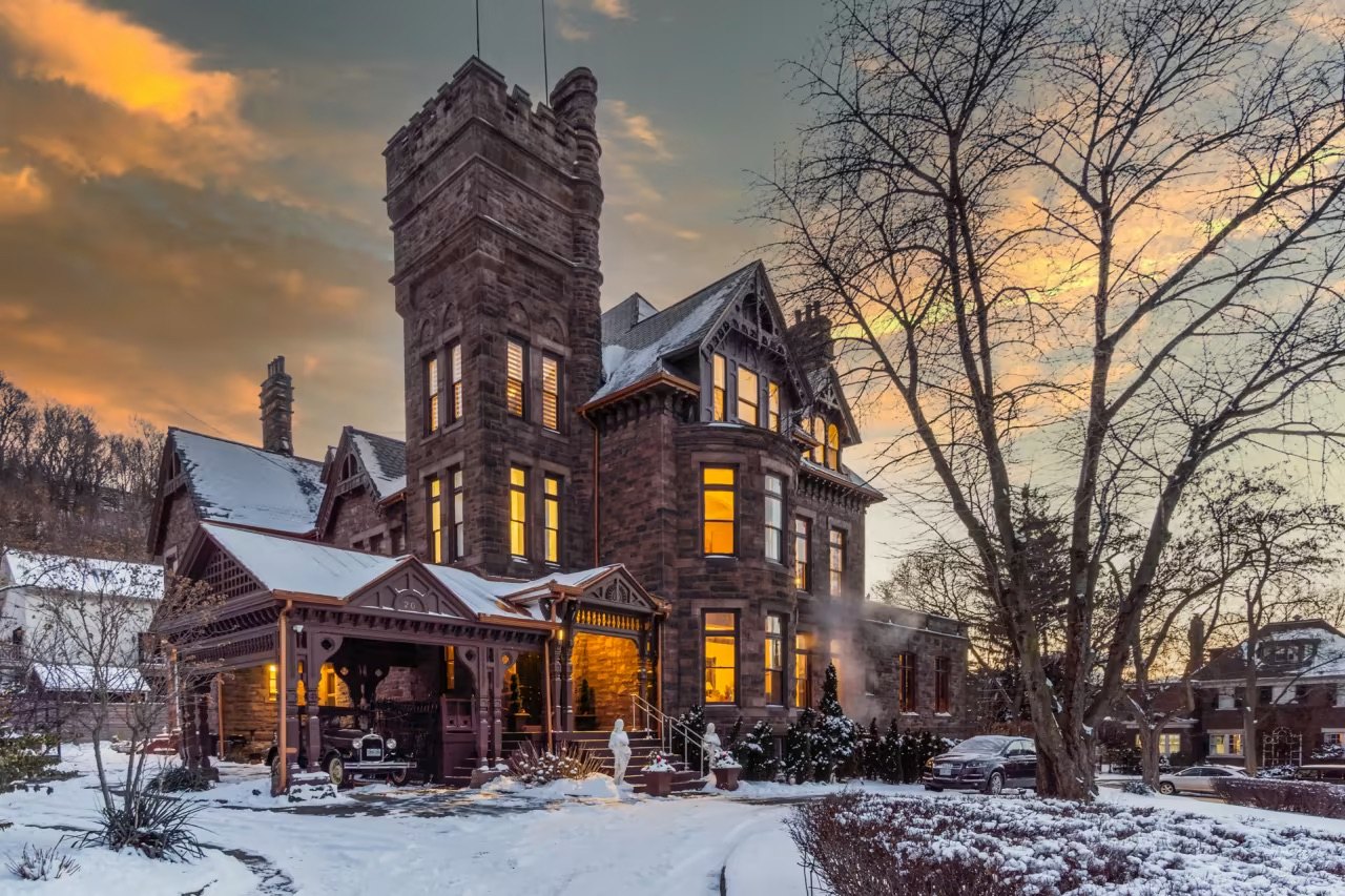 Historic stone mansion with turret and warmly lit windows in snow at sunset