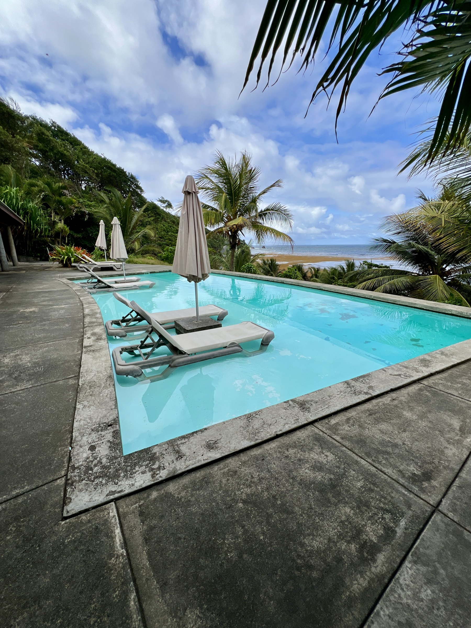 Rectangular pool with loungers and closed parasols facing a distant beach.