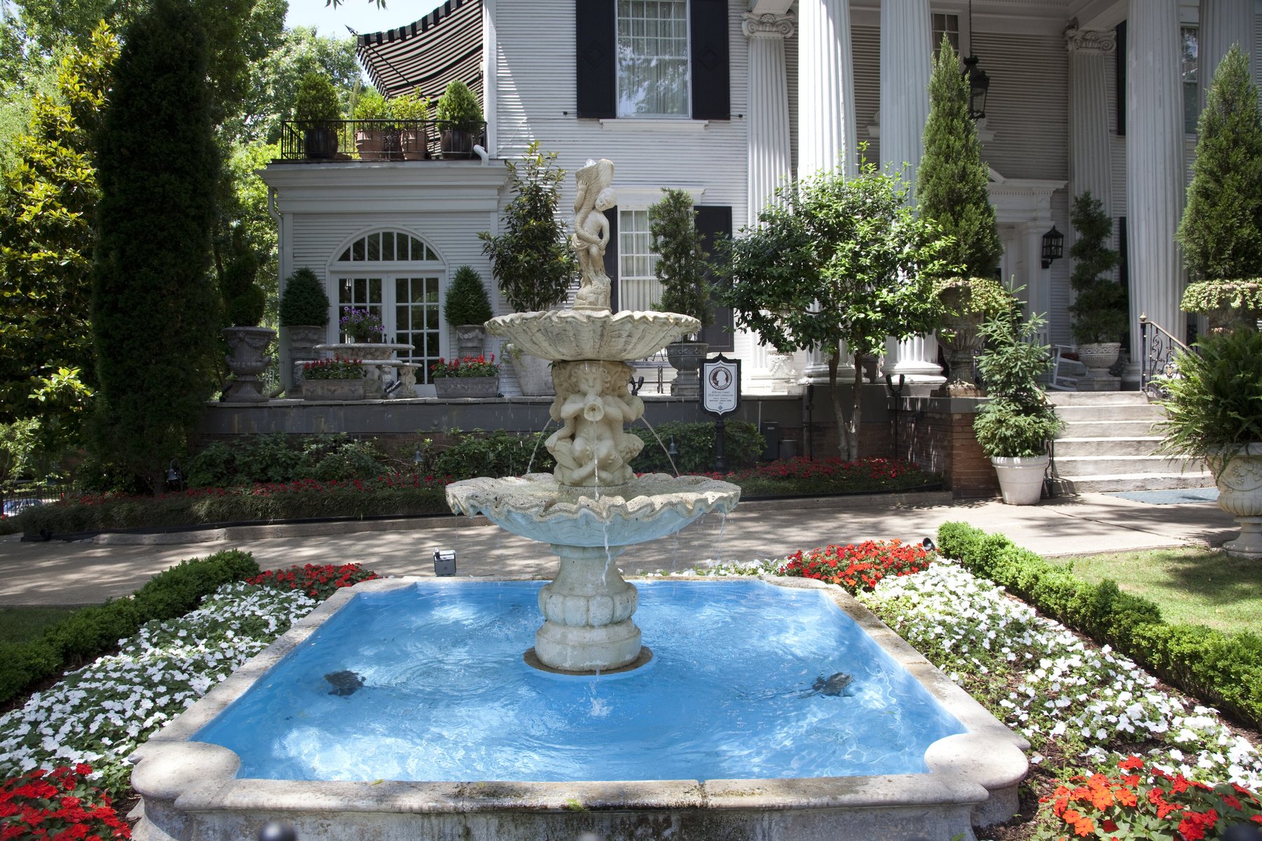 Tiered stone fountain and flower beds before a white columned mansion with black shutters