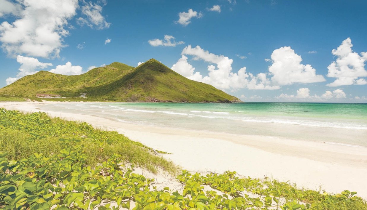Empty white sand beach curving toward a steep green headland under blue sky