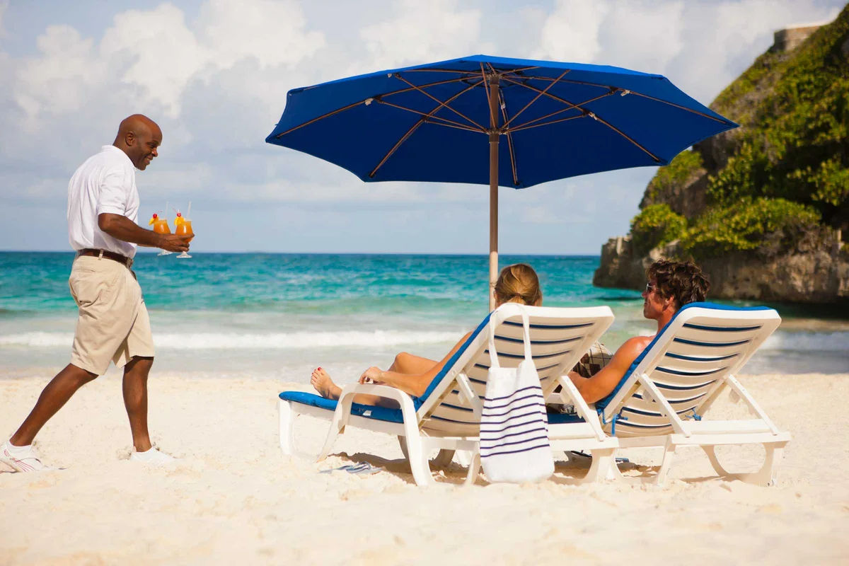 Beach service photo: a waiter in white shirt carrying two cocktails across the sand to a couple reclining on