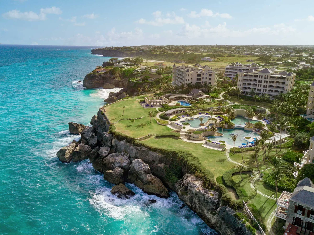Aerial photo along the cliff edge: eroded rock stacks and white surf in the foreground, clifftop lawn, the