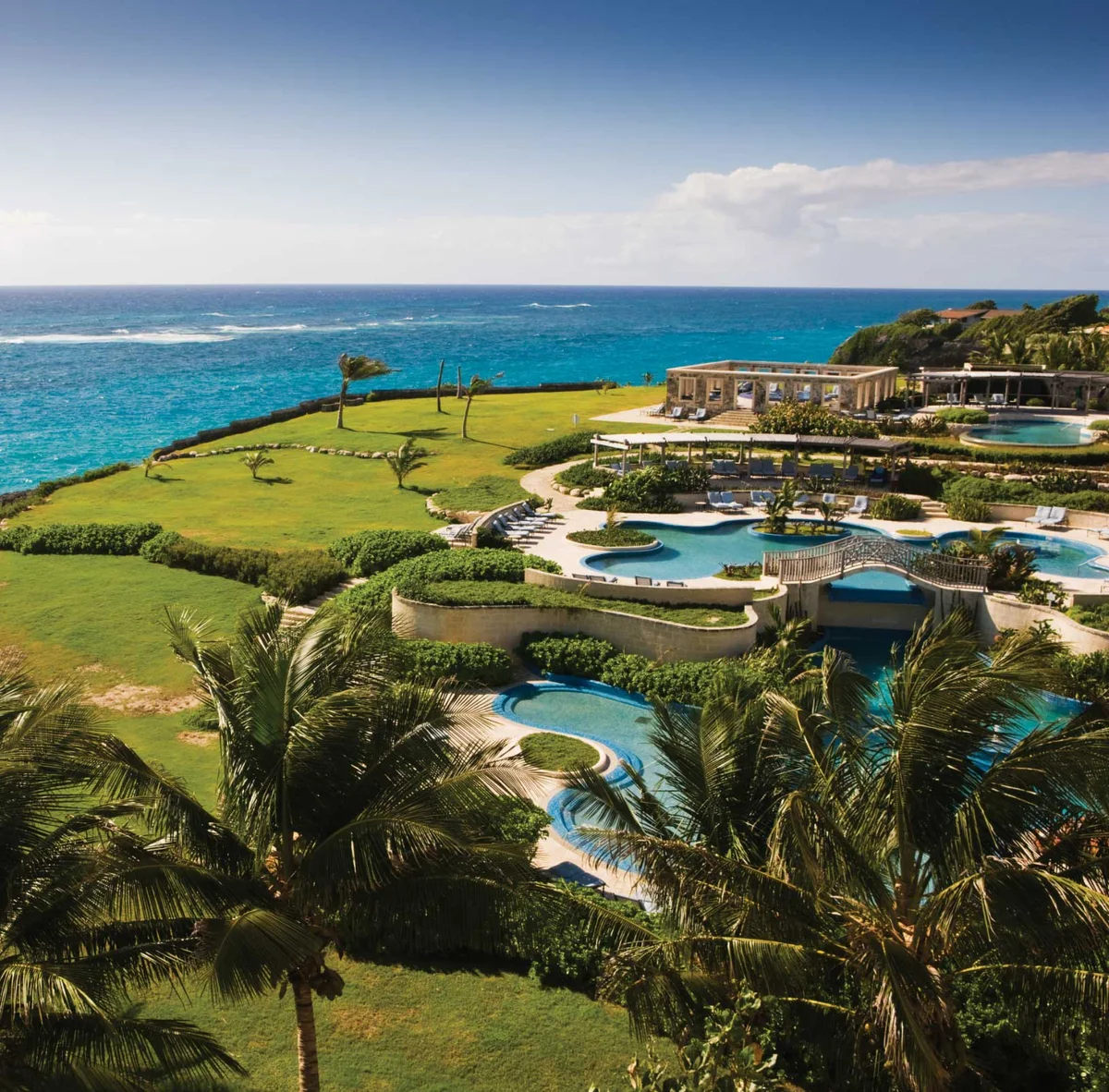 Near-square elevated photo of the terraced resort pools, stone bridge and clipped garden terraces, clifftop