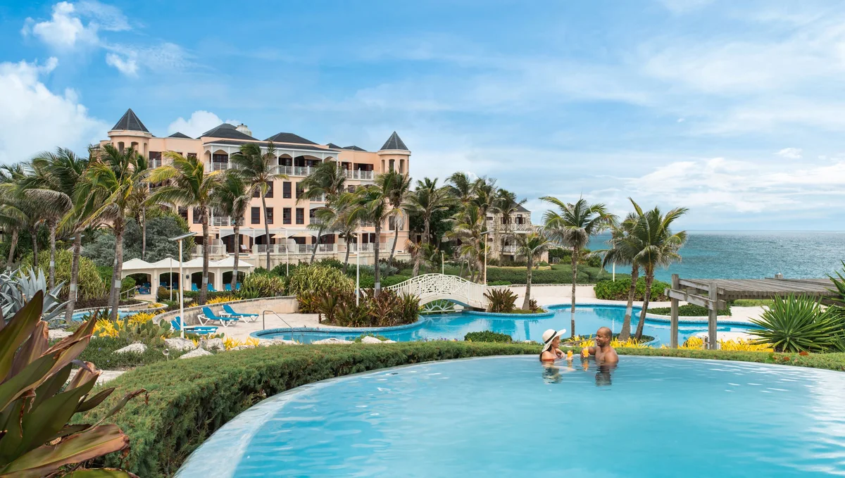 Resort pools with a couple standing in the water in the foreground, stone footbridge mid-frame, coral-stone