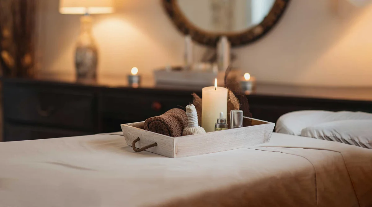 Spa treatment room detail: white wooden tray on the massage table holding a lit pillar candle, rolled brown