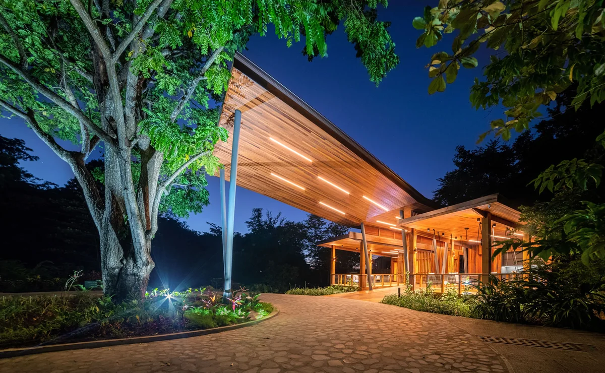 PHOTO β resort restaurant/reception pavilion lit at dusk beside a large spreading tree, cobbled forecourt. 1600x986