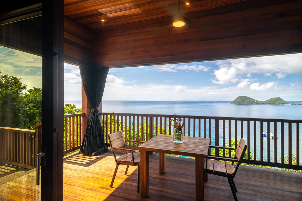 PHOTO β covered deck with timber dining table and chairs, railing, wide ocean view to an offshore island. 1600x1067