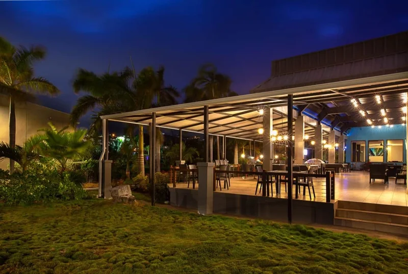A covered outdoor dining terrace at the Royal St Kitts lit at dusk, with tables under a glass canopy, palms alongside and lawn in the foreground