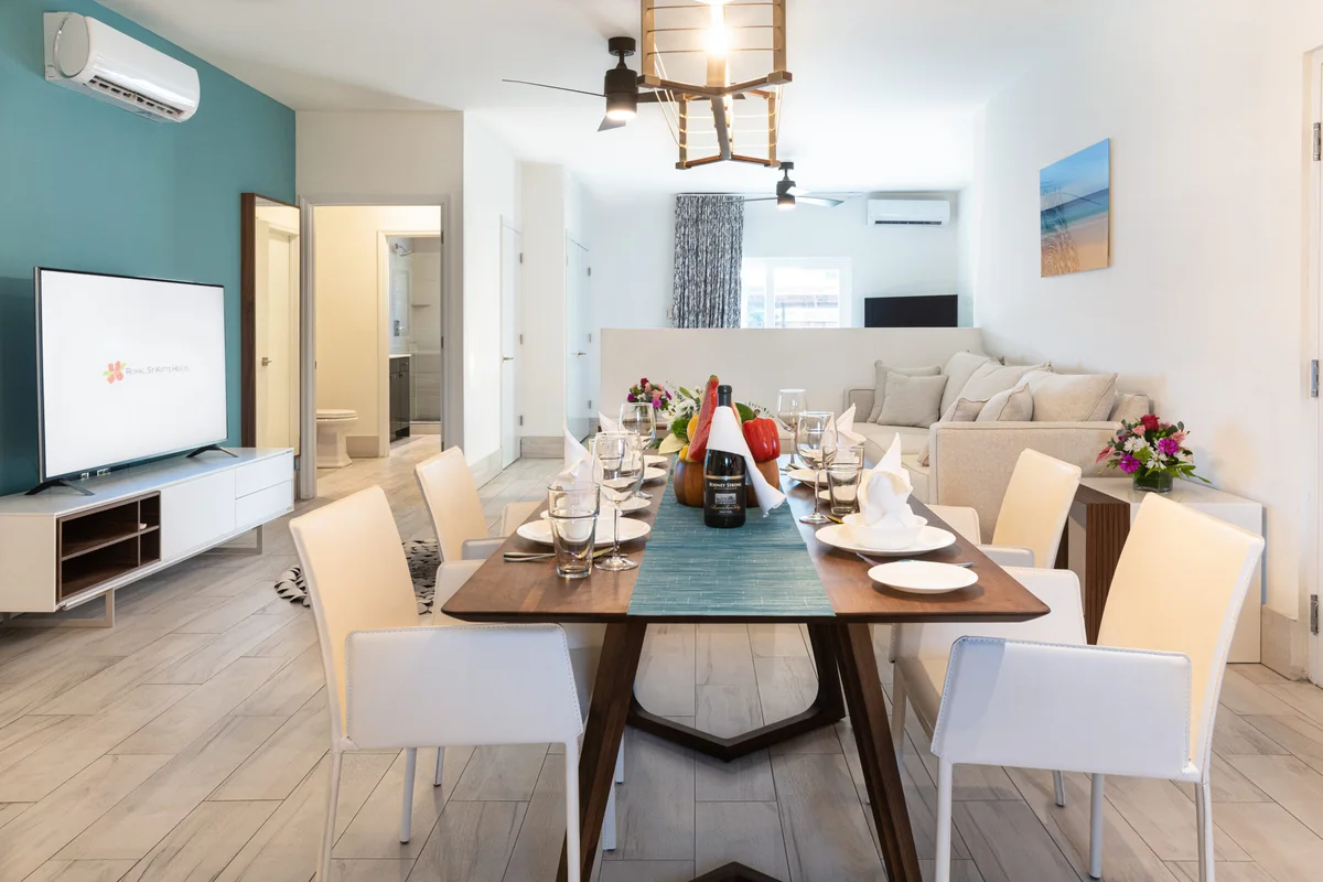 Open-plan living/dining room, long walnut table set for eight, sofa, teal accent wall; wall TV displays the