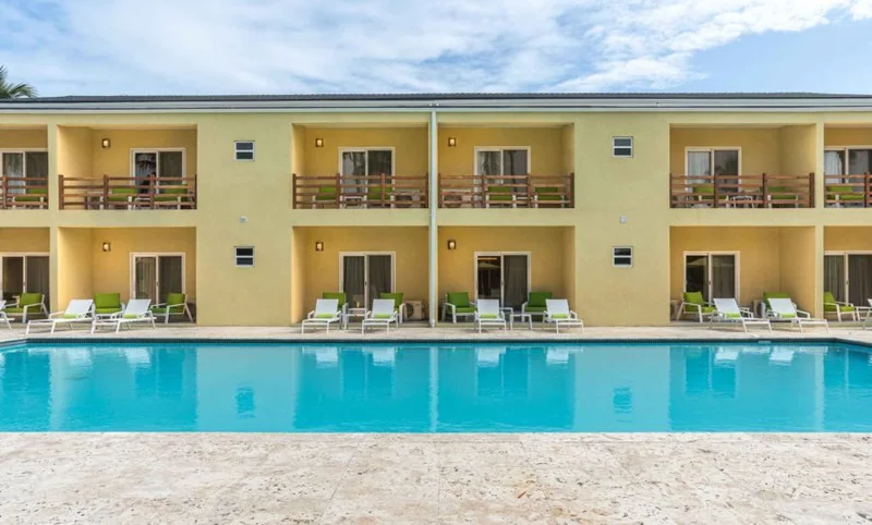 Frontal view across the pool to the two-storey yellow condo block, green loungers lined along the travertine deck