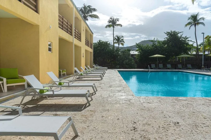 Row of white loungers with green towels along the poolside walkway beside the yellow condo building; hills behind
