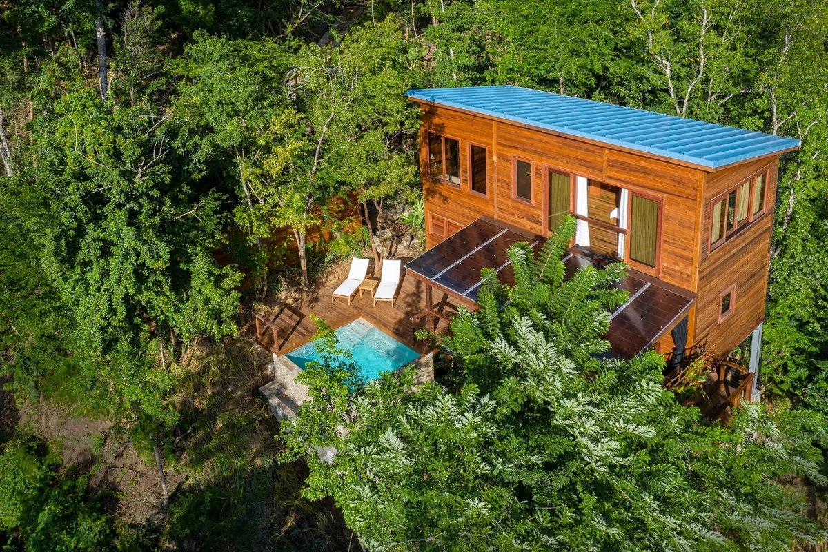 PHOTO — drone view of a timber villa with blue roof, plunge pool and sun loungers surrounded by rainforest.