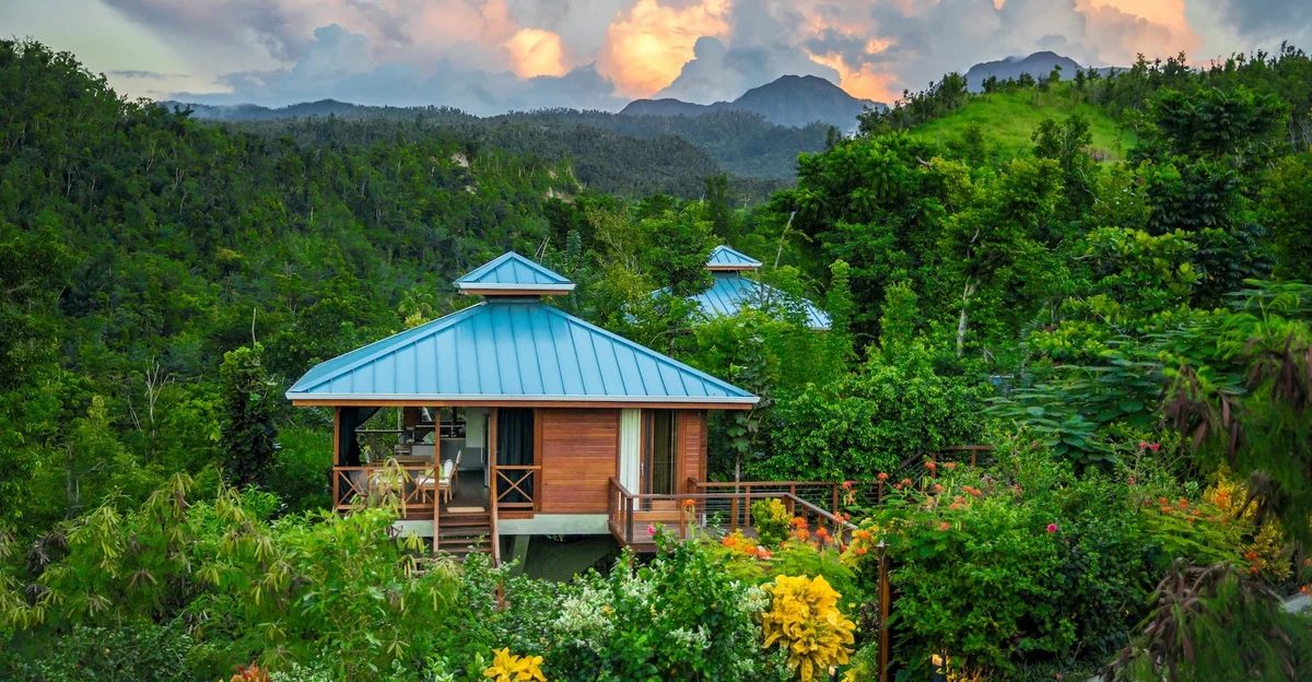 PHOTO — blue-roofed timber villa in dense rainforest at sunset, mountain peaks and dramatic sky behind. 1960x1020