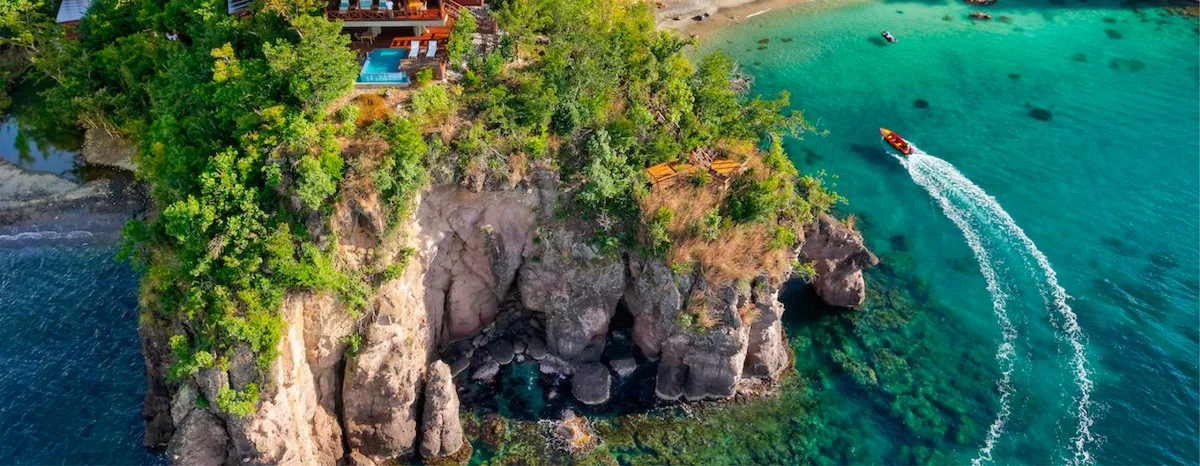 PHOTO — wide panoramic aerial banner of the headland and sea stack with a boat on turquoise water. Very