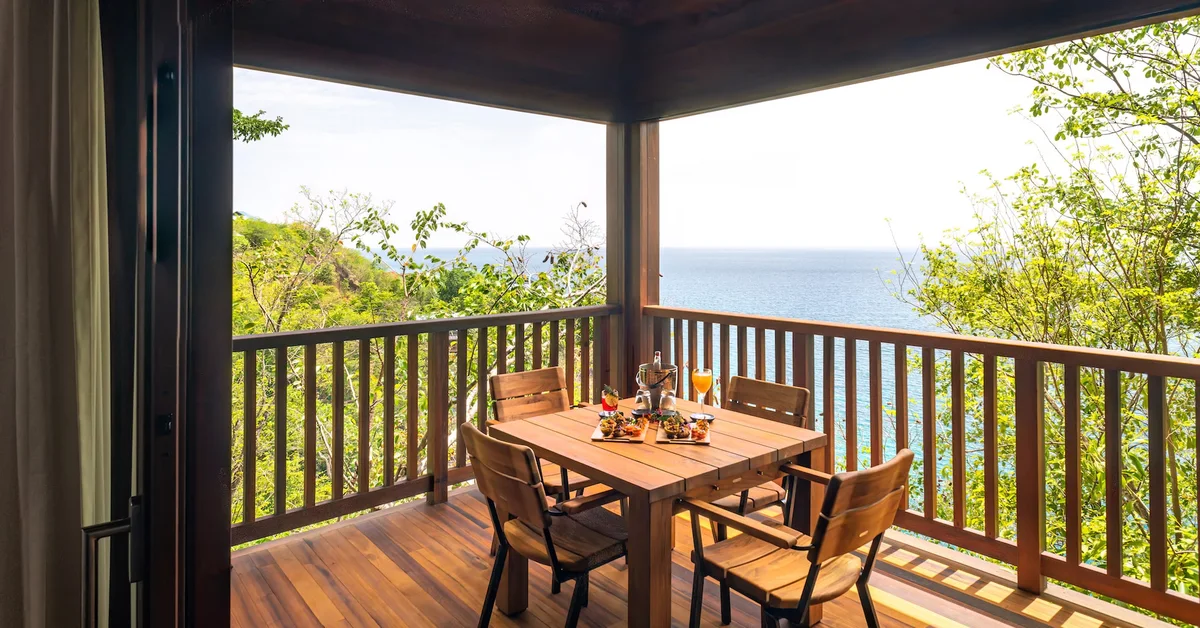 PHOTO — covered deck with timber dining table set for two, railing, sea view through the opening. 1960x1026